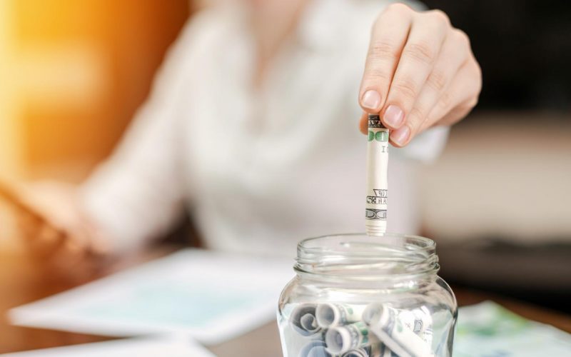 woman-dropping-banknotes-into-jar-with-rolled-banknotes-table-papers-table woman-dropping-banknotes-into-jar-with-rolled-banknotes-table-papers-table